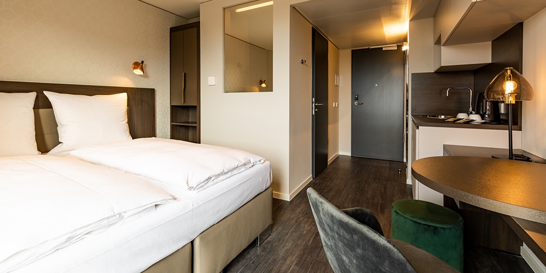 Hotelzimmer mit Doppelbett, einer Küchenzeile, einem grauen Stuhl und einem grünen Hocker and einem runden Tisch mit leuchtender Tischlampe