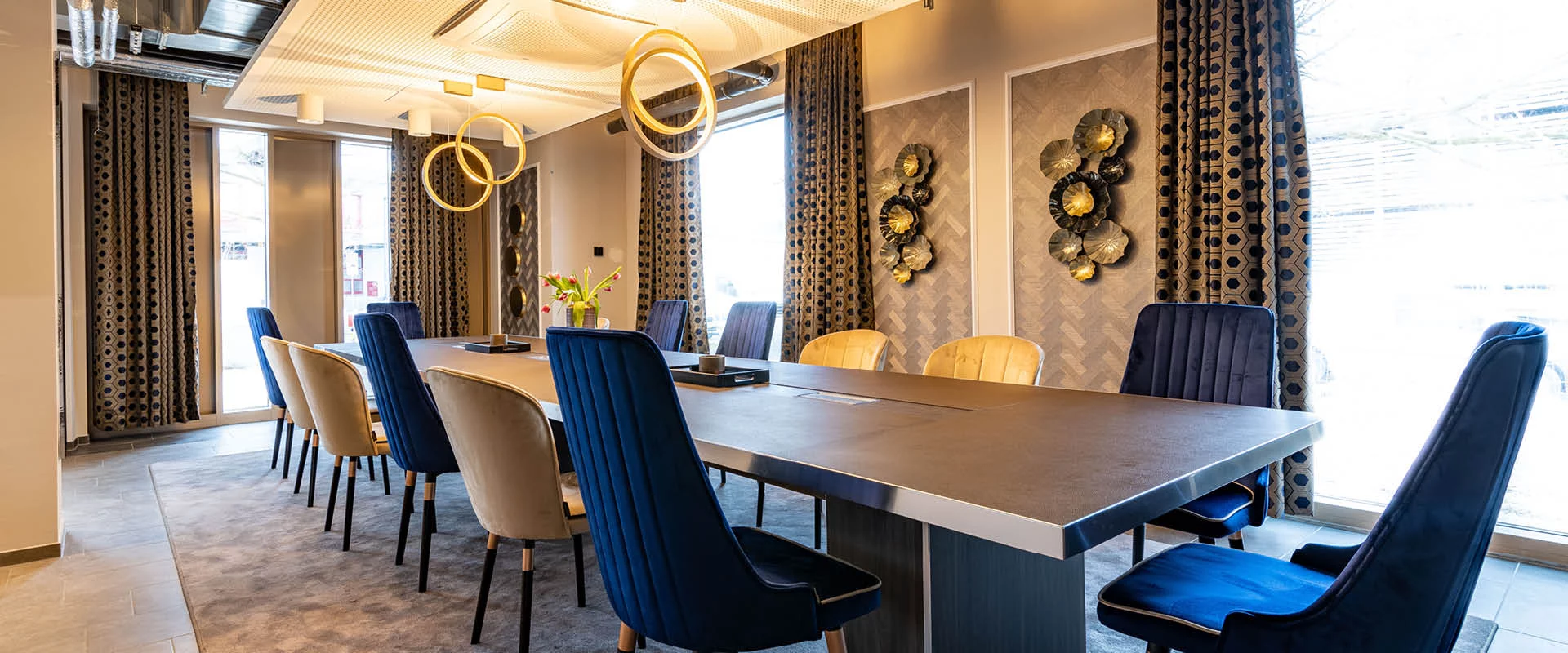 Ein Konferenzraum mit dunkelblauen und beigen Stühlen an einem langen dunklen Tisch, dekoriert mit Kerzen und einer Vase mit Blumen, sowie beige Vorhänge mit schwarzen Punkten und einem grauen Teppich.